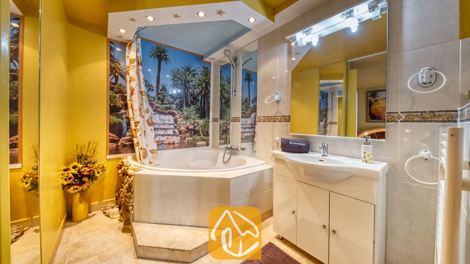 Vakantievilla's Costa Brava Spanje - Villa Geolouk - Badkamer met een hoekbad voorzien van een tropische watervalmuurschildering, een witte wastafel met een grote spiegel en gele muren.