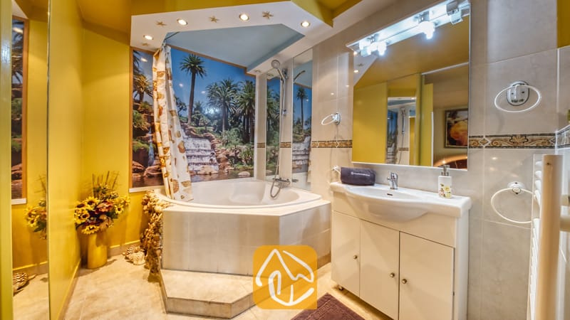 Villas de vacances Costa Brava Espagne - Villa Geolouk - Salle de bains comprenant une baignoire d'angle ornée d'une fresque murale représentant une cascade tropicale, un meuble-lavabo blanc surmonté d'un grand miroir, et des murs jaunes.