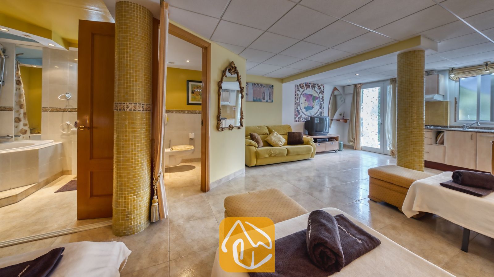 Vakantievilla's Costa Brava Spanje - Villa Geolouk - Het ruime interieur van Villa Geolouk, voorzien van plavuizen vloeren, gele zuilen, een zitgedeelte met een bank en bedden met donkerbruine handdoeken op de voorgrond.