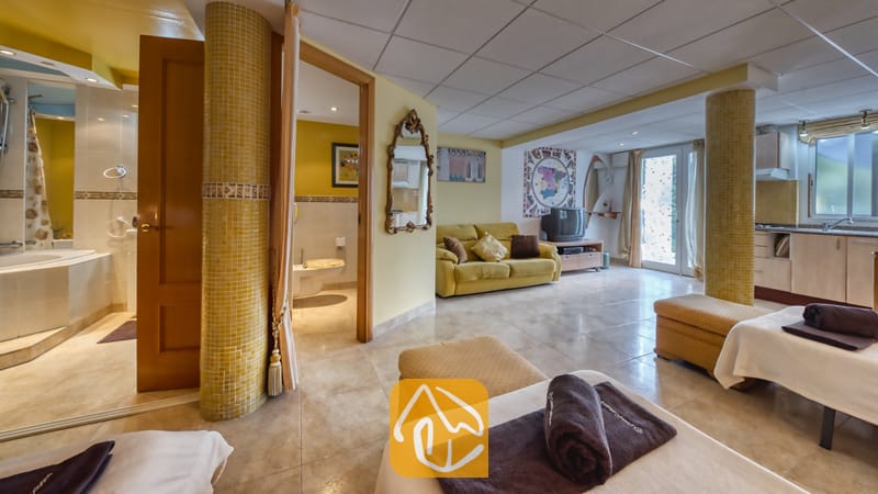 Vakantievilla's Costa Brava Spanje - Villa Geolouk - Het ruime interieur van Villa Geolouk, voorzien van plavuizen vloeren, gele zuilen, een zitgedeelte met een bank en bedden met donkerbruine handdoeken op de voorgrond.