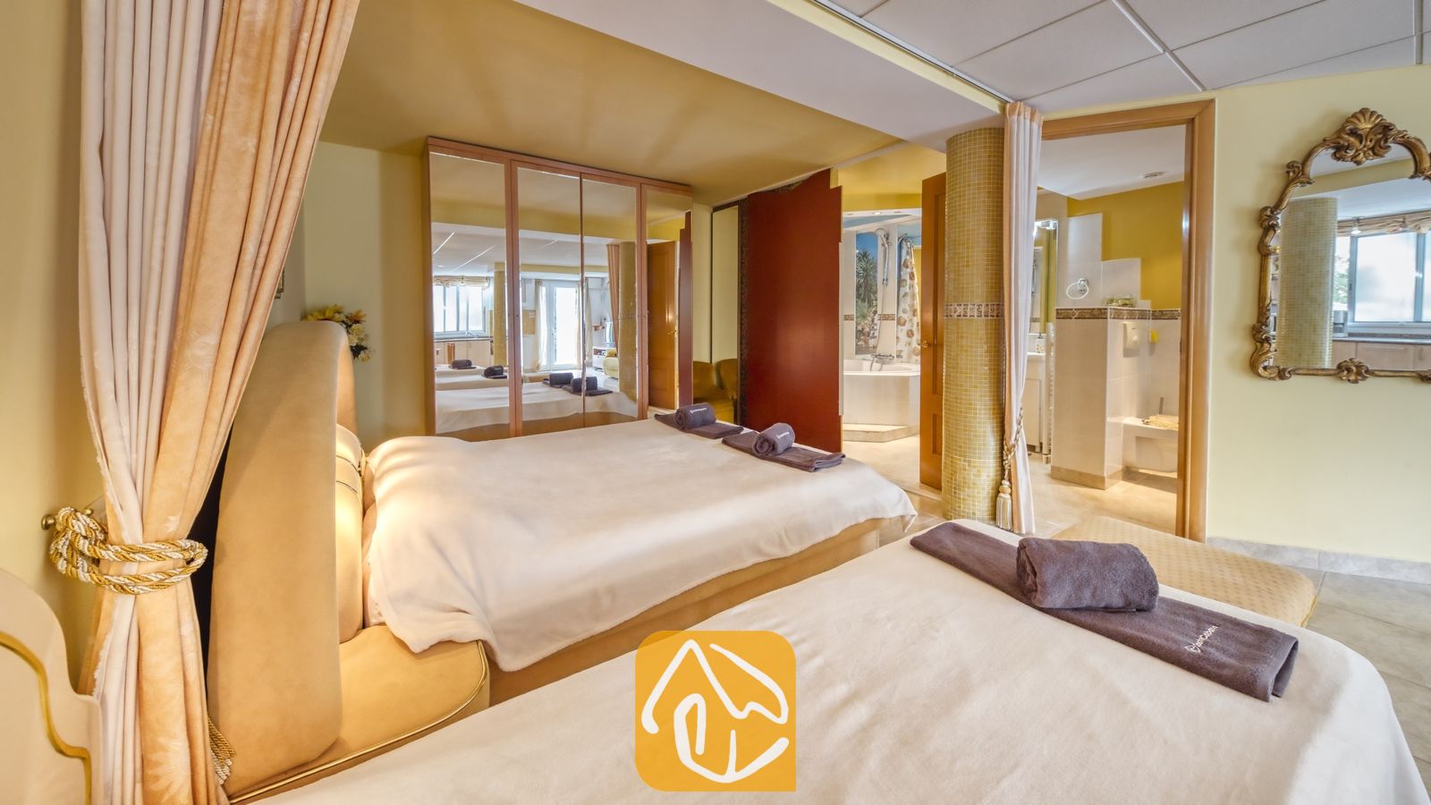 Vakantievilla's Costa Brava Spanje - Villa Geolouk - Slaapkamer in Villa Geolouk met twee bedden voorzien van wit beddengoed en opgerolde grijze handdoeken, grote spiegeldeuren van de kledingkast en een open doorgang naar een en-suite badkamer.