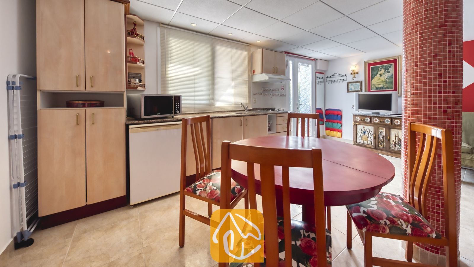 Vakantievilla's Costa Brava Spanje - Villa Geolouk - Ronde rode houten eettafel met stoelen voorzien van bloemkussens in de keuken van Villa Geolouk, voorzien van lichte houten kasten en een rode mozaïekzuil.