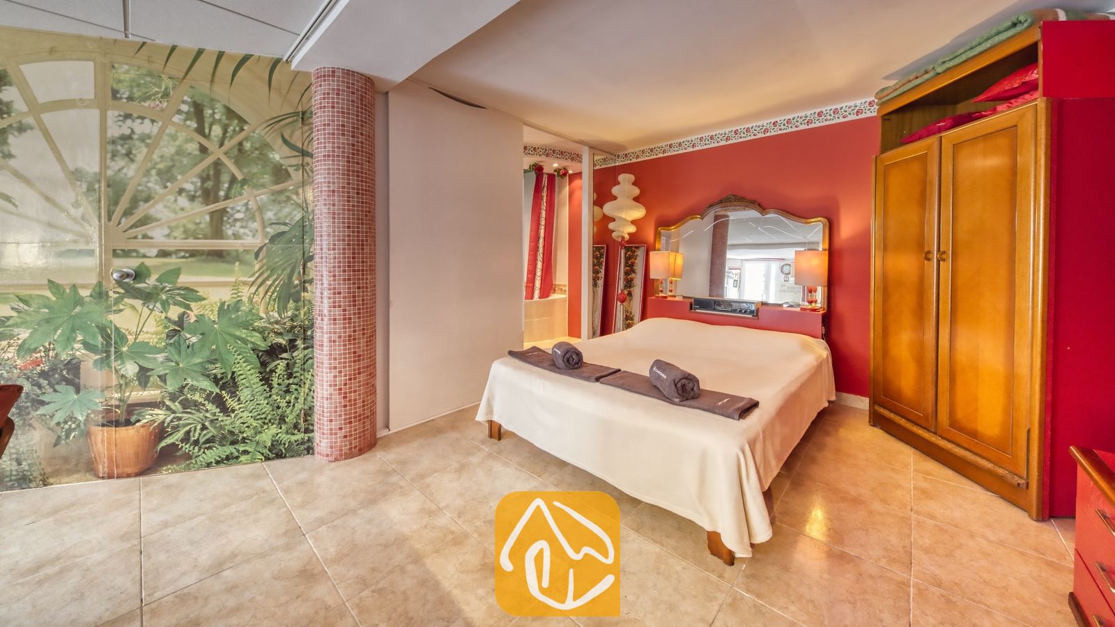 Vakantievilla's Costa Brava Spanje - Villa Geolouk - Slaapkamer met rode muren, een tweepersoonsbed met opgerolde handdoeken, een grote houten kledingkast, tegelvloer en een muurschildering met een plantenmotief.
