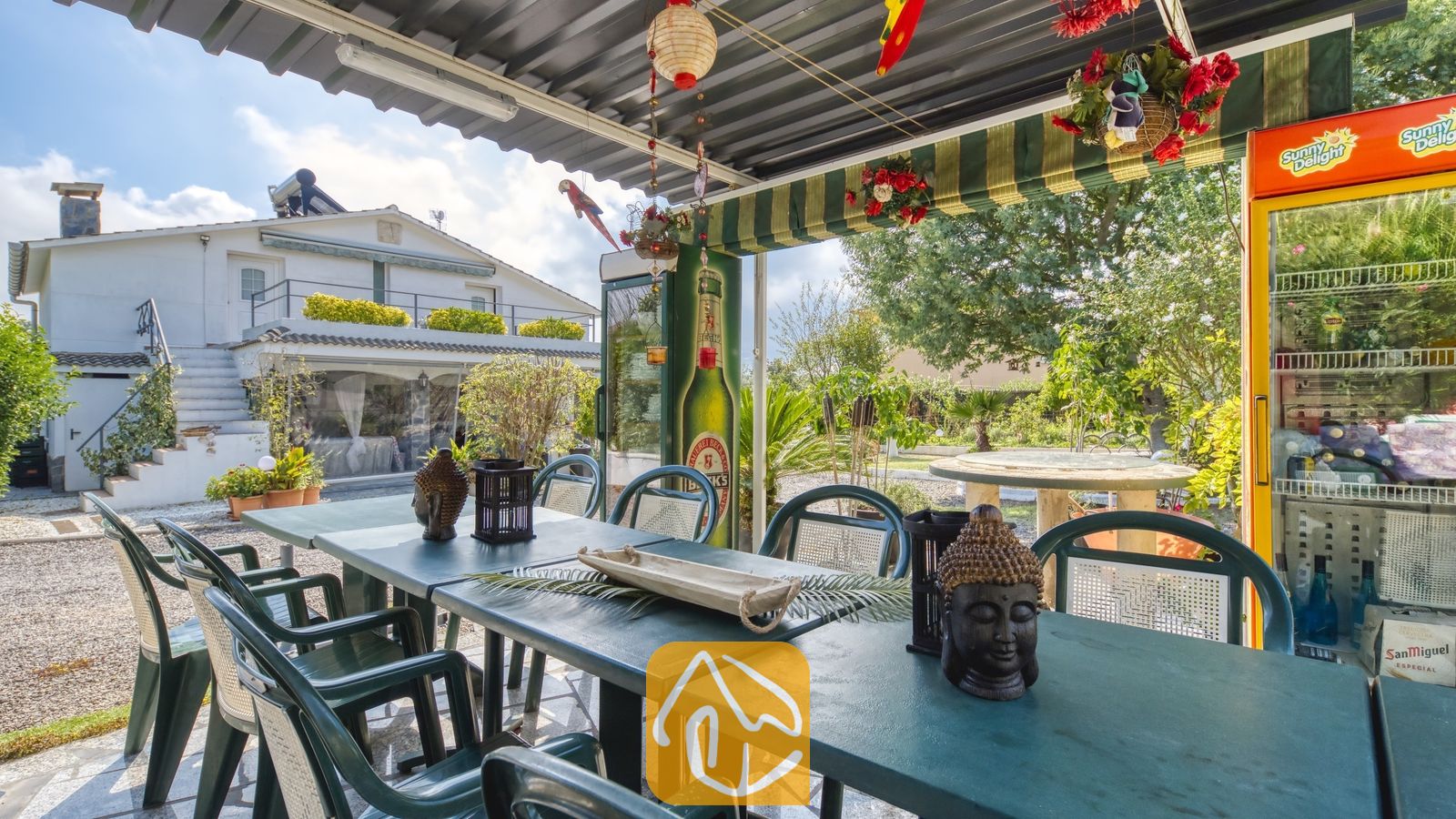 Vakantievilla's Costa Brava Spanje - Villa Geolouk - Overdekt buitenterras bij Villa Geolouk met donkergroene eettafels, bijpassende stoelen, decoratie in de vorm van een Boeddhahoofd en een koelkast.