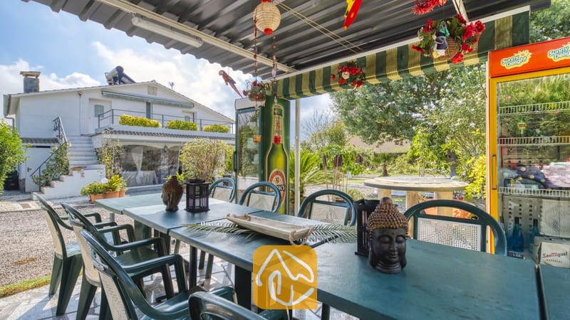 Vakantievilla's Costa Brava Spanje - Villa Geolouk - Overdekt buitenterras bij Villa Geolouk met donkergroene eettafels, bijpassende stoelen, decoratie in de vorm van een Boeddhahoofd en een koelkast.