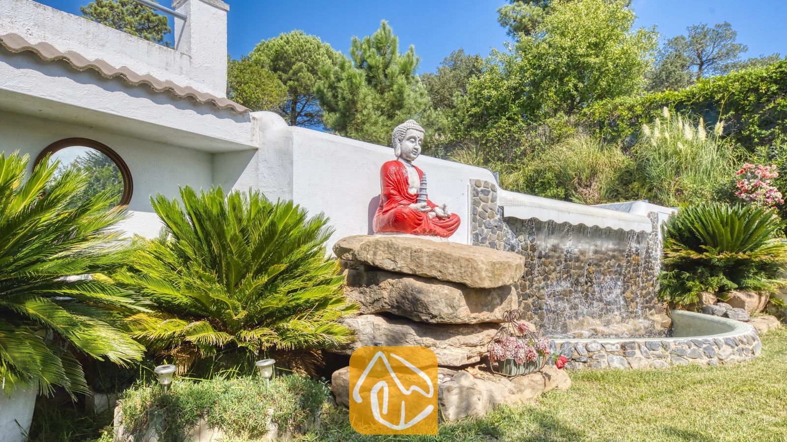 Vakantievilla's Costa Brava Spanje - Villa Geolouk - Beeld van een zittende Boeddha in rode gewaden, rustend op gestapelde stenen rotsen naast een stenen waterpartij en palmbomen bij Villa Geolouk