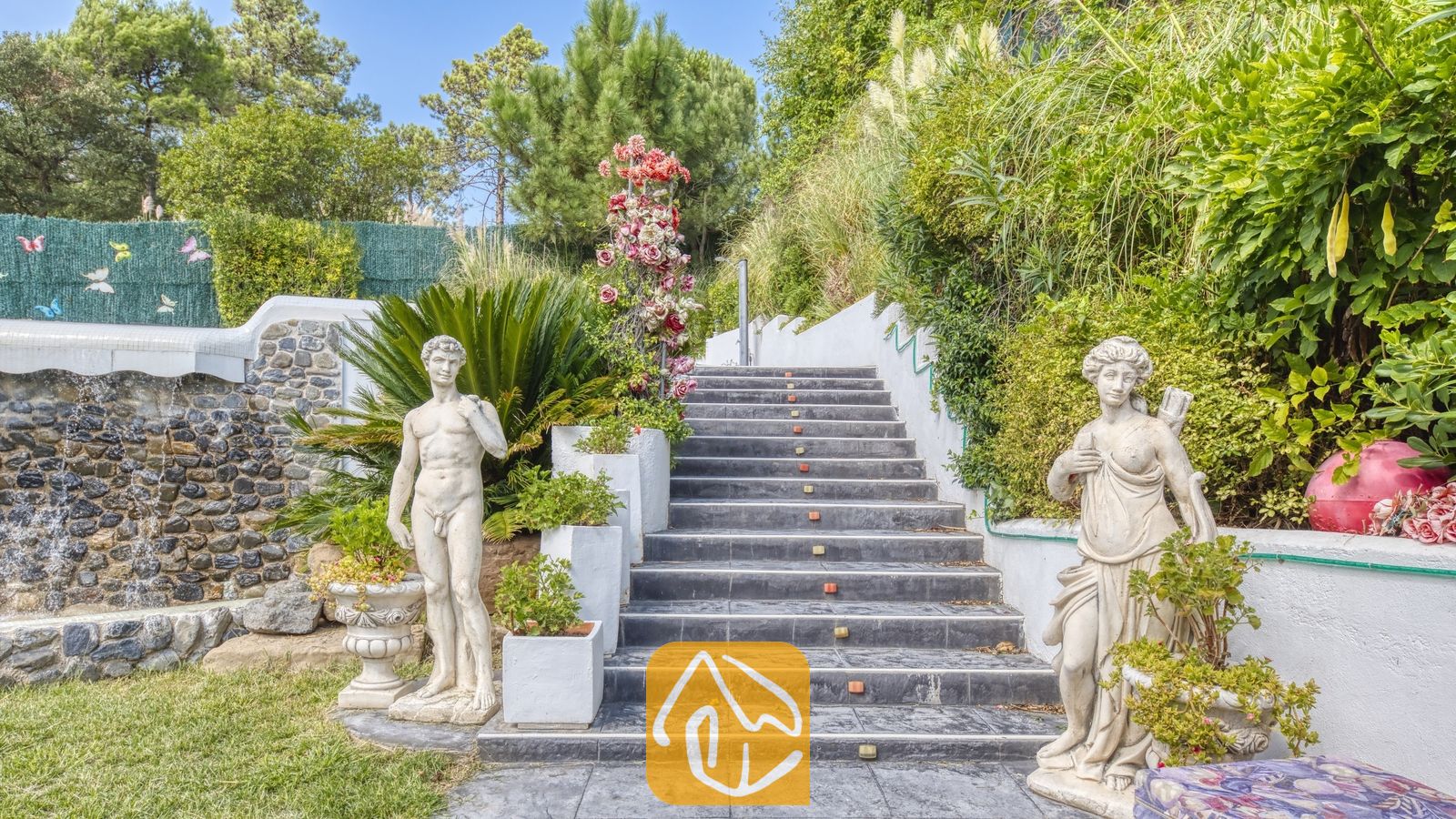 Vakantievilla's Costa Brava Spanje - Villa Geolouk - Buiten stenen trappen bij Villa Geolouk, geflankeerd door klassieke beelden, weelderig groen en een decoratieve stenen muur.