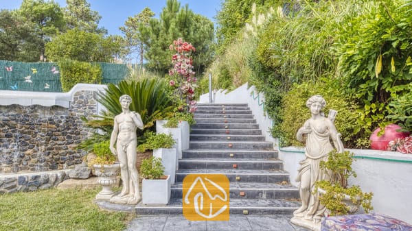Vakantievilla's Costa Brava Spanje - Villa Geolouk - Buiten stenen trappen bij Villa Geolouk, geflankeerd door klassieke beelden, weelderig groen en een decoratieve stenen muur.
