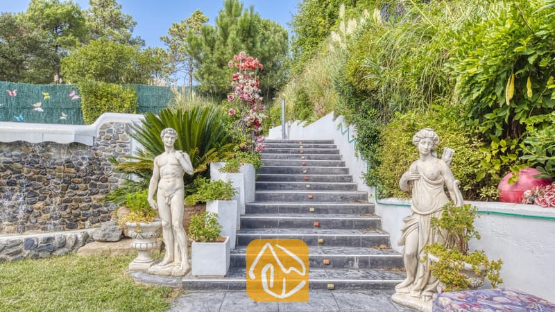 Vakantievilla's Costa Brava Spanje - Villa Geolouk - Buiten stenen trappen bij Villa Geolouk, geflankeerd door klassieke beelden, weelderig groen en een decoratieve stenen muur.
