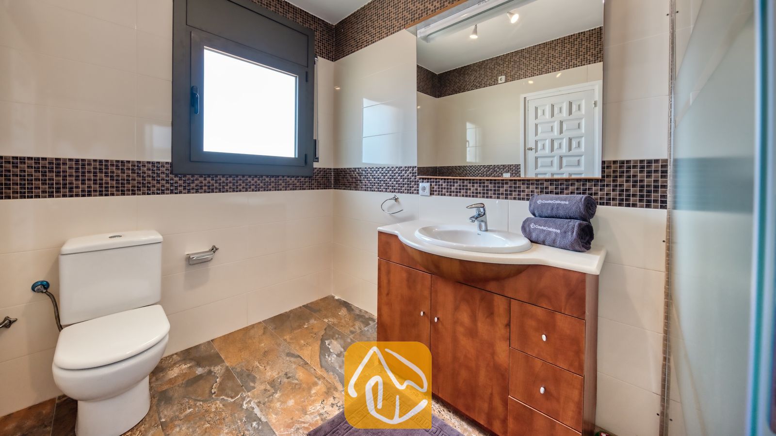 Vakantievilla's Costa Brava Spanje - Villa Jaruco - Moderne badkamer met een houten badkamermeubel, een witte keramische wastafel, een toilet en een grote spiegel omlijst door bruine mozaïektegels.