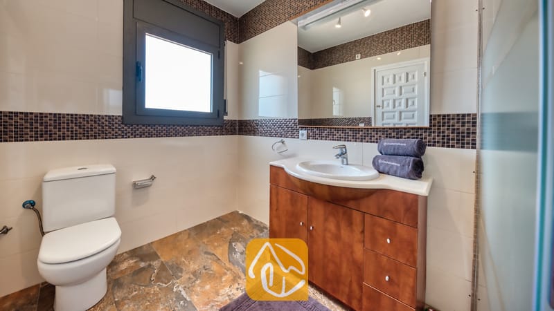Villas de vacances Costa Brava Espagne - Villa Jaruco - Salle de bains moderne comprenant un meuble coiffeuse en bois, un lavabo en céramique blanche, des toilettes et un grand miroir encadré de mosaïques marron.