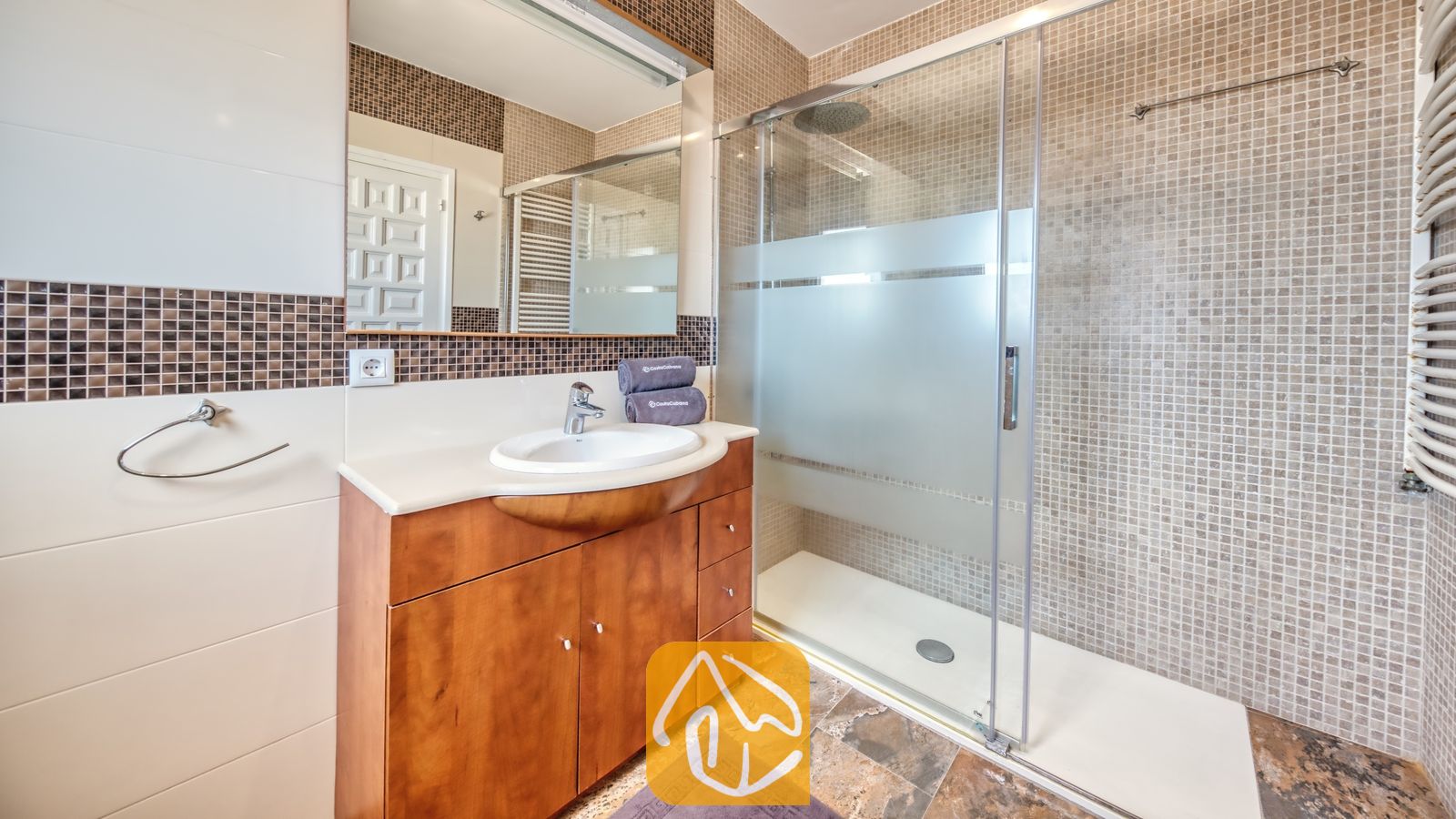 Vakantievilla's Costa Brava Spanje - Villa Jaruco - Moderne badkamer met een houten badkamermeubel, een witte keramische wastafel en een glazen inloopdouche met beige mozaïektegels.
