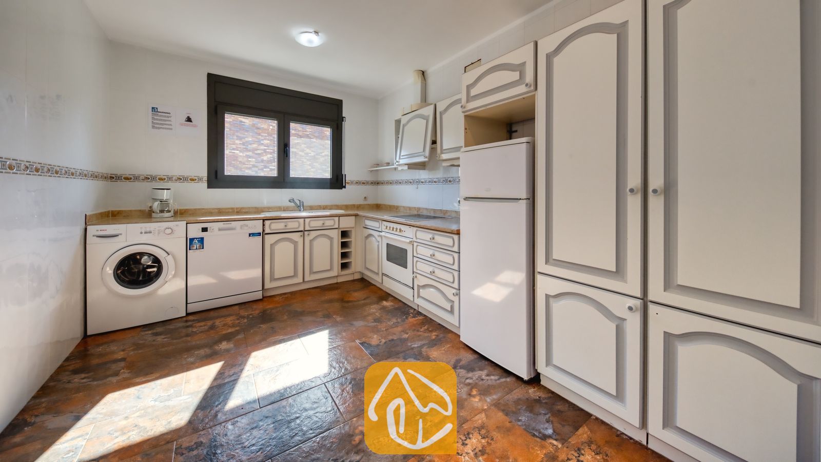 Vakantievilla's Costa Brava Spanje - Villa Jaruco - Keukeninterieur met witte kasten, donkere tegelvloeren, een wasmachine, vaatwasser en een witte koelkast.