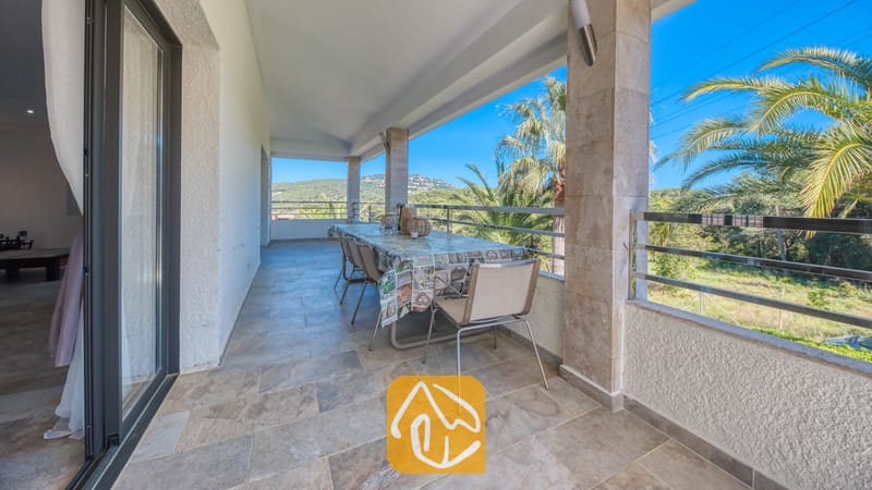 Vakantievilla's Costa Brava Spanje - Villa Jaruco - Overdekt terras bij Villa Jaruco, voorzien van een lange eettafel, tegelvloer, stenen steunpilaren en een stalen balustrade met uitzicht op palmbomen en heuvels in de verte.
