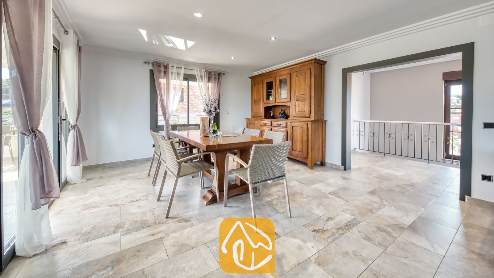 Vakantievilla's Costa Brava Spanje - Villa Jaruco - Houten eettafel, omringd door geweven grijze stoelen binnen in Villa Jaruco, voorzien van tegelvloeren en een grote houten kast tegen de wand.