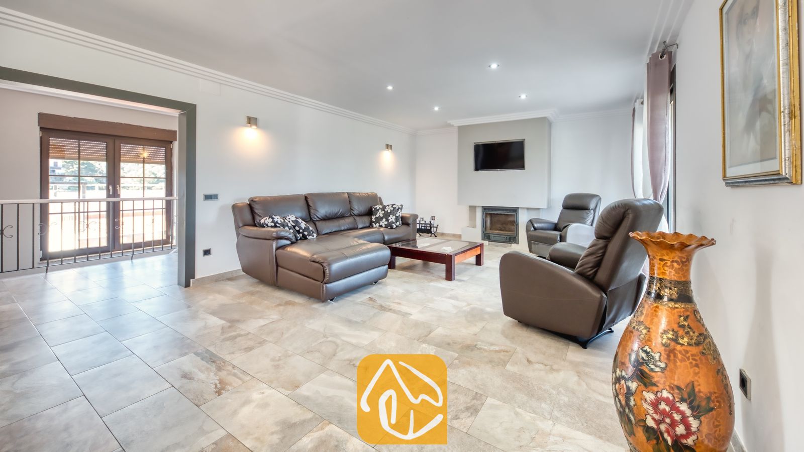 Vakantievilla's Costa Brava Spanje - Villa Jaruco - Woonkamer van Villa Jaruco met een bruine L-vormige lederen bank, bijpassende fauteuils, een open haard onder een aan de muur gemonteerde tv en een tegelvloer.