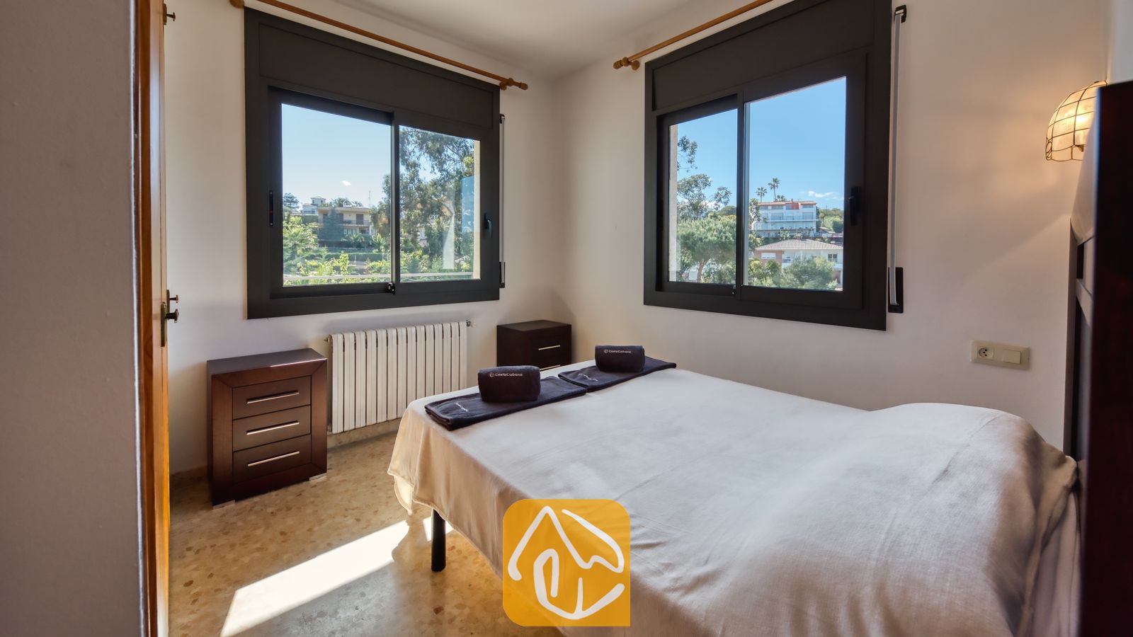 Vakantievilla's Costa Brava Spanje - Villa Jaruco - Slaapkamer met een tweepersoonsbed opgemaakt met wit beddengoed, een donkerhouten ladekast en twee ramen met uitzicht op het groen buiten.