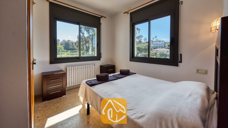 Villas de vacances Costa Brava Espagne - Villa Jaruco - Chambre avec lit double habillé de linge de blanc, commode en bois foncé et deux fenêtres offrant une vue sur la verdure extérieure.