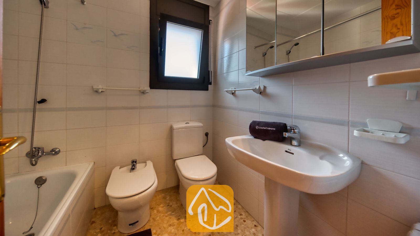 Vakantievilla's Costa Brava Spanje - Villa Jaruco - Wit betegelde badkamer met een vrijstaande wastafel, toilet, bidet en een ligbad onder een raam met een donker kozijn.