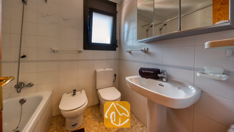 Villas de vacances Costa Brava Espagne - Villa Jaruco - Salle de bains carrelée de blanc comprenant un lavabo sur pied, des toilettes, un bidet et une baignoire sous une fenêtre à cadre foncé.