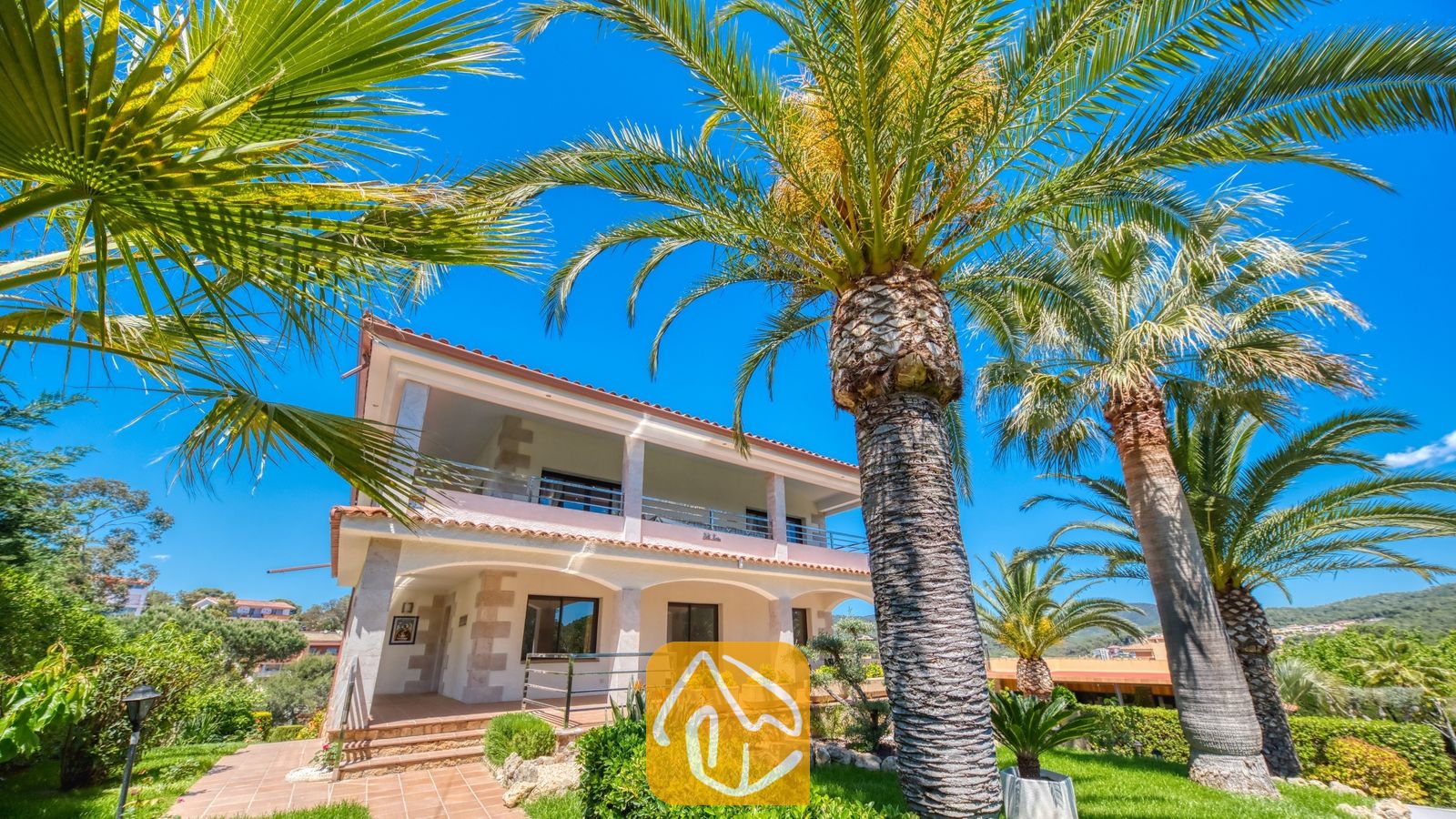 Vakantievilla's Costa Brava Spanje - Villa Jaruco - Buitenaanzicht van Villa Jaruco, omlijst door hoge palmbomen tegen een helderblauwe lucht, met zicht op een huis van twee verdiepingen met een overdekt terras met bogen beneden en een balkon boven.