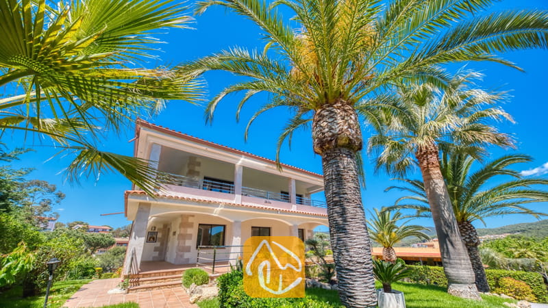 Vakantievilla's Costa Brava Spanje - Villa Jaruco - Buitenaanzicht van Villa Jaruco, omlijst door hoge palmbomen tegen een helderblauwe lucht, met zicht op een huis van twee verdiepingen met een overdekt terras met bogen beneden en een balkon boven.