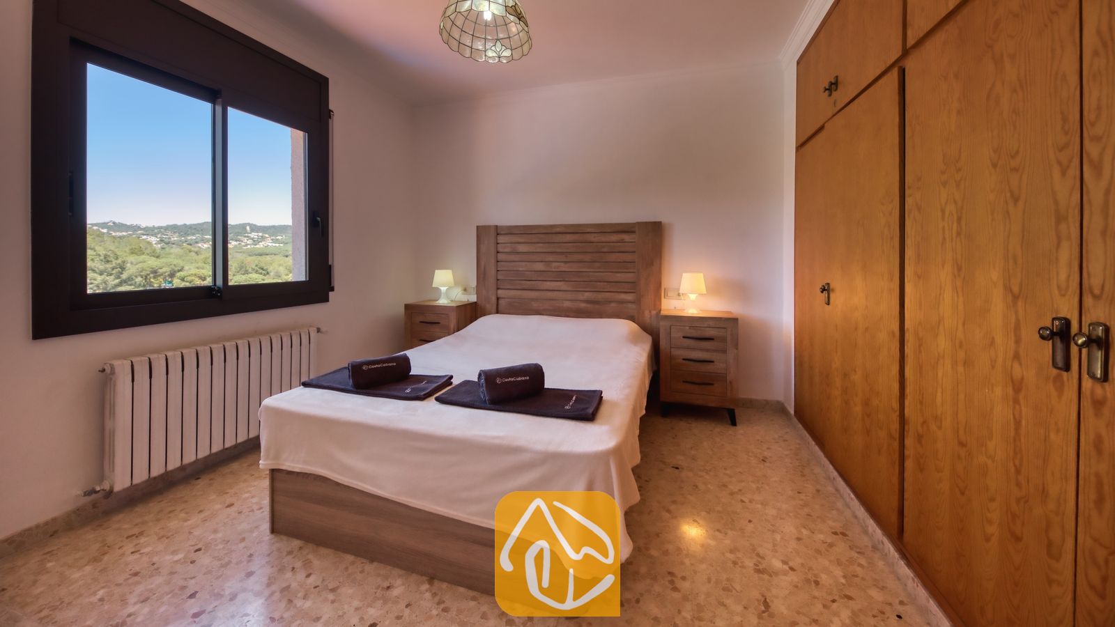 Vakantievilla's Costa Brava Spanje - Villa Jaruco - Slaapkamer met een tweepersoonsbed, een houten hoofdbord, twee nachtkastjes met lampen, een groot raam met uitzicht op groene heuvels en ingebouwde houten kasten.