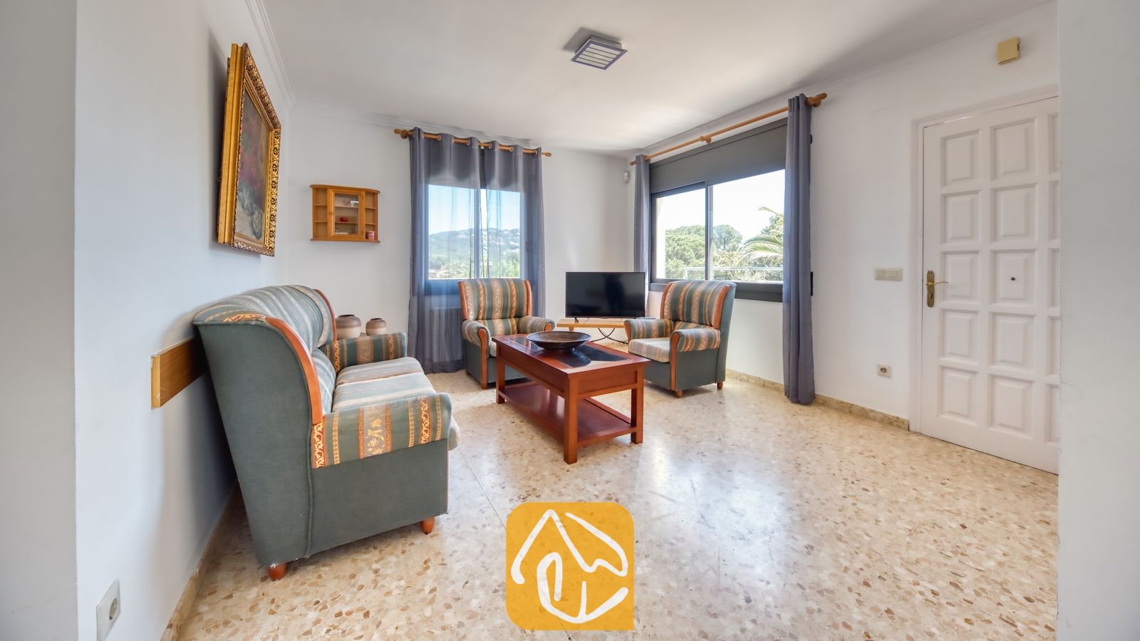 Vakantievilla's Costa Brava Spanje - Villa Jaruco - Woonkamer met een stoffen bank met patroon en bijbehorende fauteuils, een houten salontafel, tegelvloer en twee ramen met blauwe gordijnen bij Villa Jaruco.