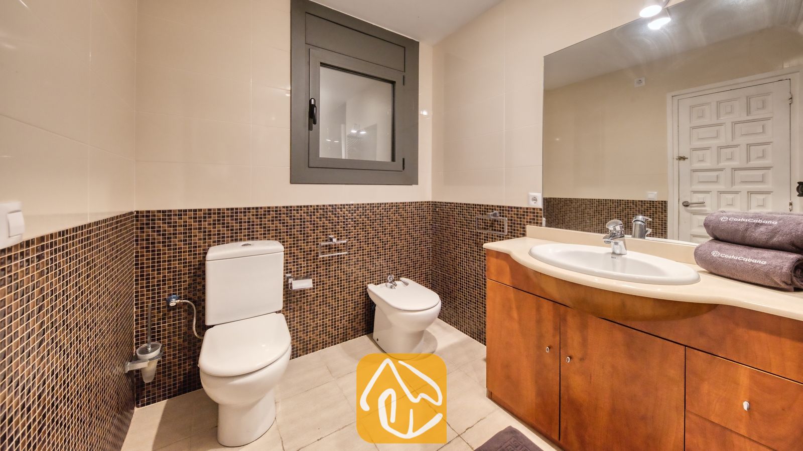 Vakantievilla's Costa Brava Spanje - Villa Jaruco - Badkamer voorzien van een wit toilet, bidet en een houten badkamermeubel met een geïntegreerde wasbak onder een grote spiegel en muren van bruine mozaïektegels.