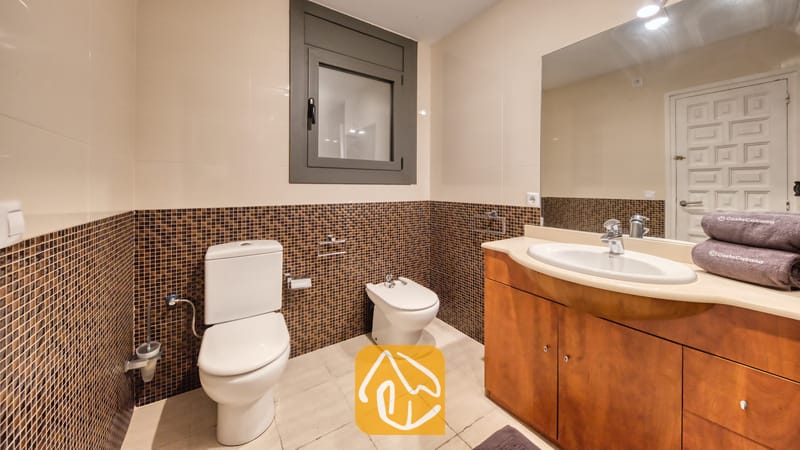 Villas de vacances Costa Brava Espagne - Villa Jaruco - Salle de bains comprenant des toilettes blanches, un bidet et un meuble sous-vasque en bois avec vasque intégrée sous un grand miroir et des murs en mosaïque marron.