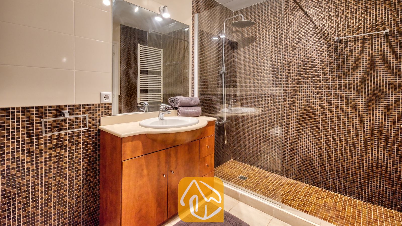 Vakantievilla's Costa Brava Spanje - Villa Jaruco - Moderne badkamer met muren van bruine mozaïektegels, een houten badkamermeubel met een witte waskom en een glazen inloopdouche.