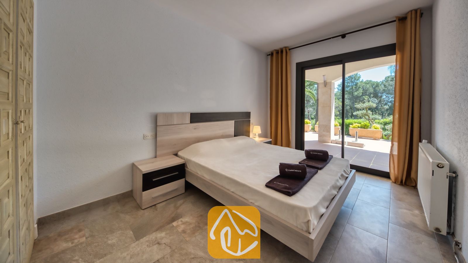 Vakantievilla's Costa Brava Spanje - Villa Jaruco - Slaapkamer met een tweepersoonsbed, bijpassende houten nachtkastjes en een glazen schuifpui die uitkomt op een verhard terras met groen uitzicht.