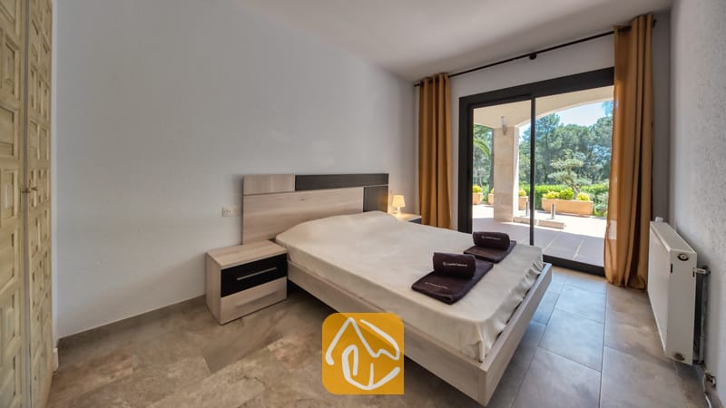Villas de vacances Costa Brava Espagne - Villa Jaruco - Chambre avec lit double, tables de chevet en bois assorties et porte-fenêtre coulissante s'ouvrant sur une terrasse pavée agrémentée de verdure extérieure.