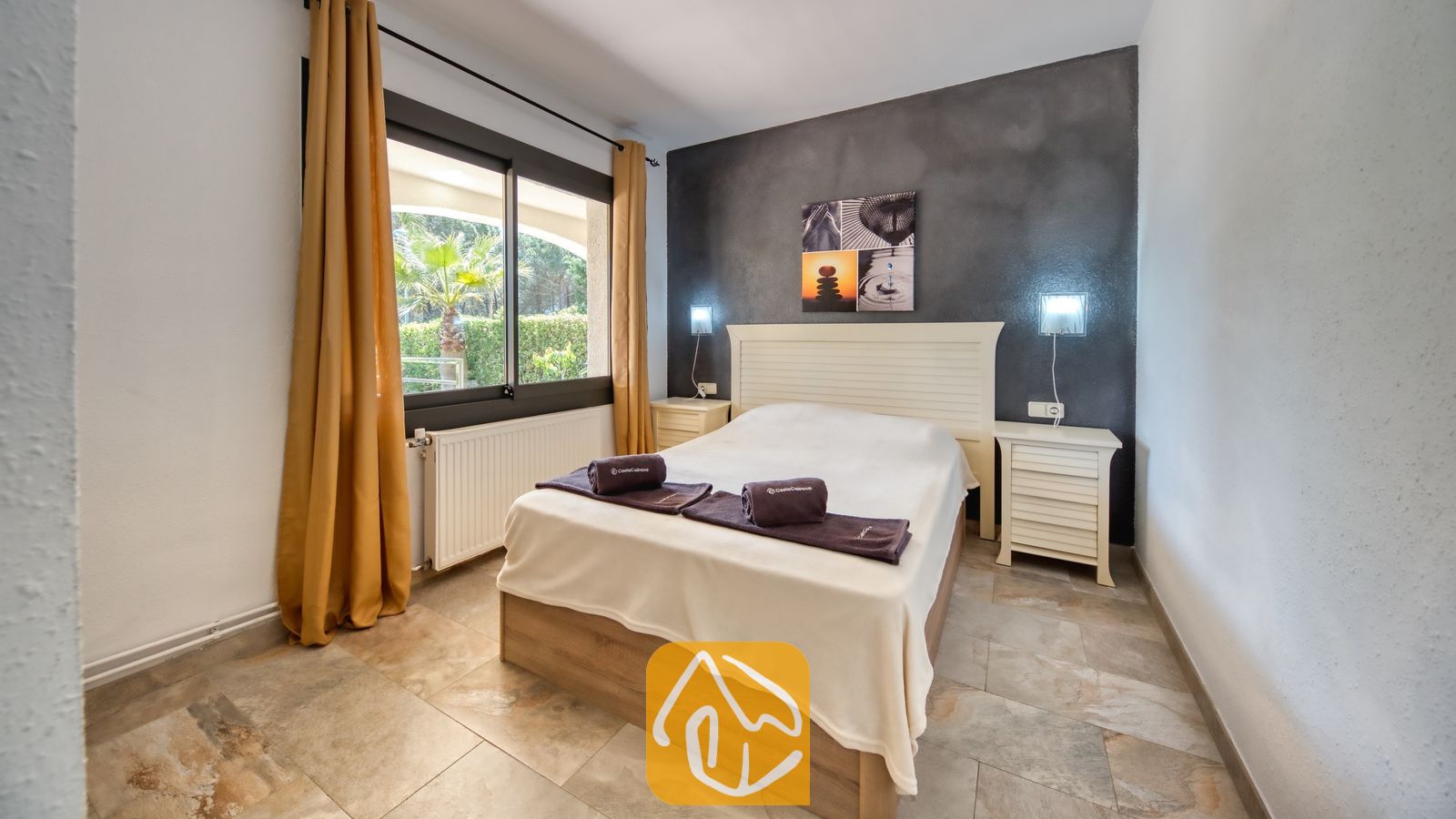 Vakantievilla's Costa Brava Spanje - Villa Jaruco - Slaapkamer met een wit bed, een donkergrijze accentmuur, bijpassende witte nachtkastjes en gele gordijnen die een raam omlijsten met uitzicht op buitenbegroeiing.