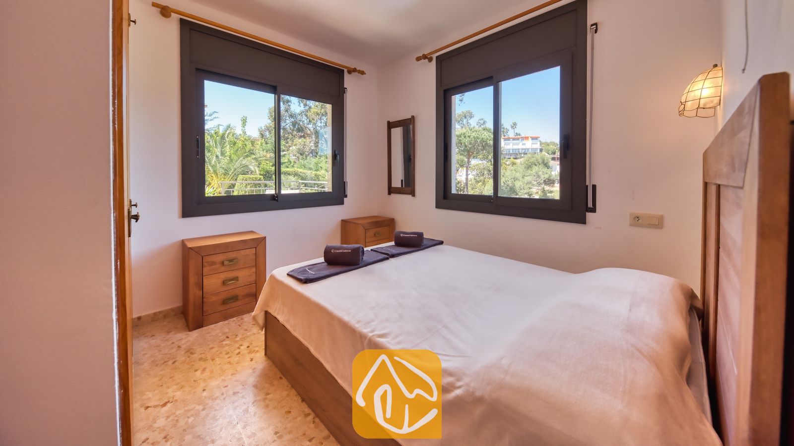 Vakantievilla's Costa Brava Spanje - Villa Jaruco - Slaapkamer met houten bedframe, tegelvloer en twee ramen met uitzicht op het buitengroen.