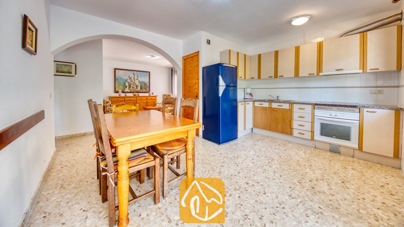 Vakantievilla's Costa Brava Spanje - Villa Jaruco - Houten eettafel met gestoffeerde stoelen in Villa Jaruco, met een aangrenzende keuken voorzien van lichte houten kasten en een helderblauwe koelkast.