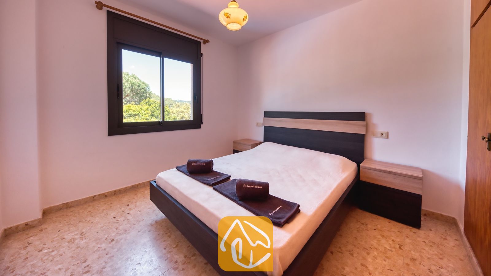 Vakantievilla's Costa Brava Spanje - Villa Jaruco - Slaapkamer met een tweepersoonsbed, een donker houten hoofdbord, twee nachtkastjes, opgerolde handdoeken, tegelvloer en een raam met uitzicht op groene bomen buiten.