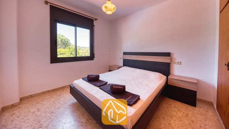 Villas de vacances Costa Brava Espagne - Villa Jaruco - Chambre avec un lit double, une tête de lit en bois sombre, deux tables de chevet, des serviettes enroulées, un sol carrelé et une fenêtre offrant une vue sur les arbres verdoyants à l'extérieur.