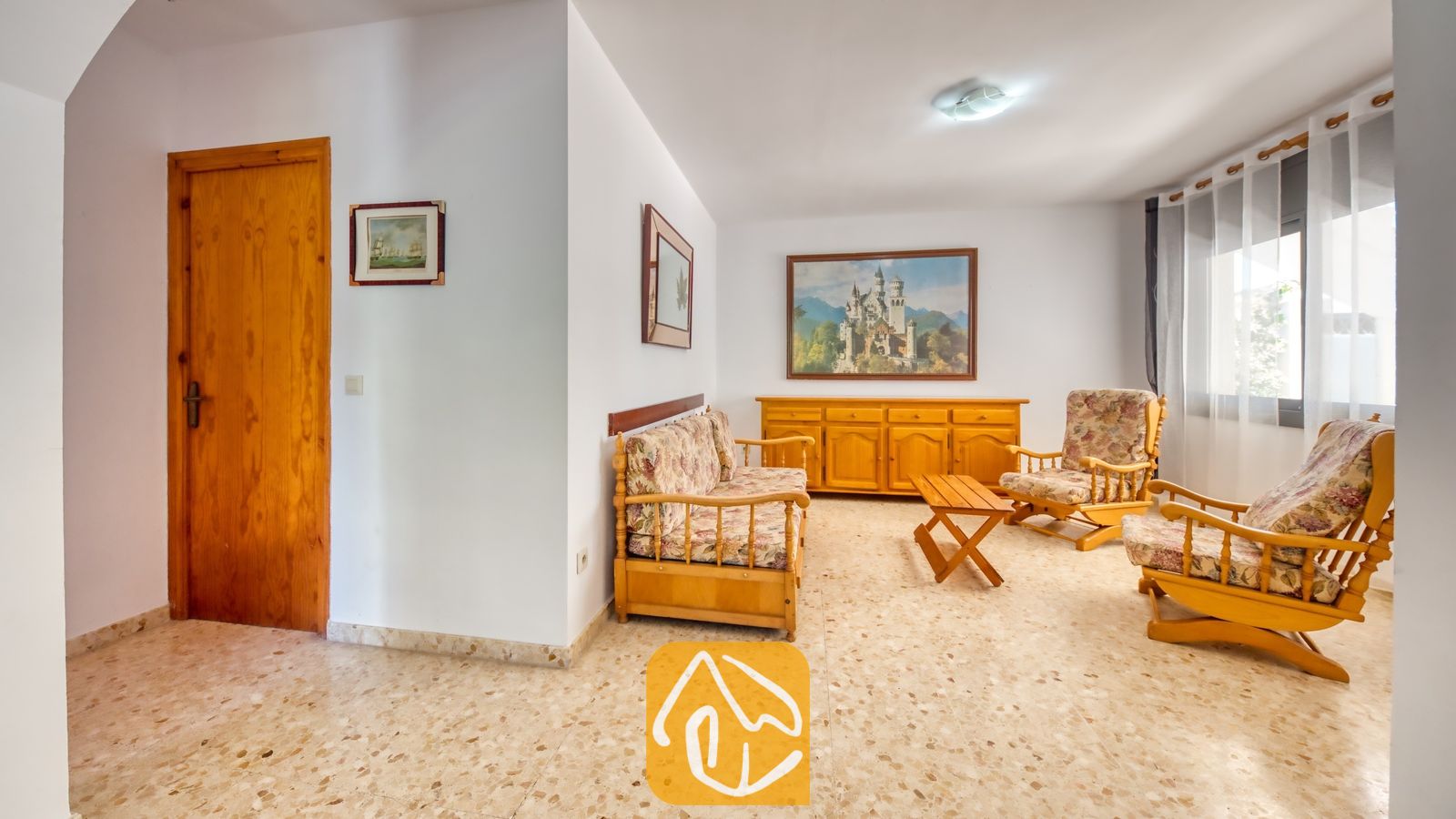 Vakantievilla's Costa Brava Spanje - Villa Jaruco - Woonkamer van Villa Jaruco met houten meubels voorzien van kussens met bloemenprint, een tegelvloer en wanddecoratie met een landschapsthema.