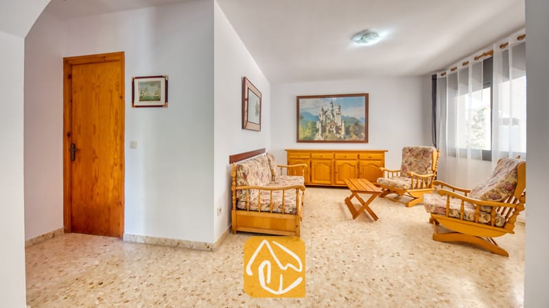 Villas de vacances Costa Brava Espagne - Villa Jaruco - Séjour de la Villa Jaruco doté de mobilier en bois avec des coussins à motifs floraux, d'un sol carrelé et de tableaux représentant des paysages.