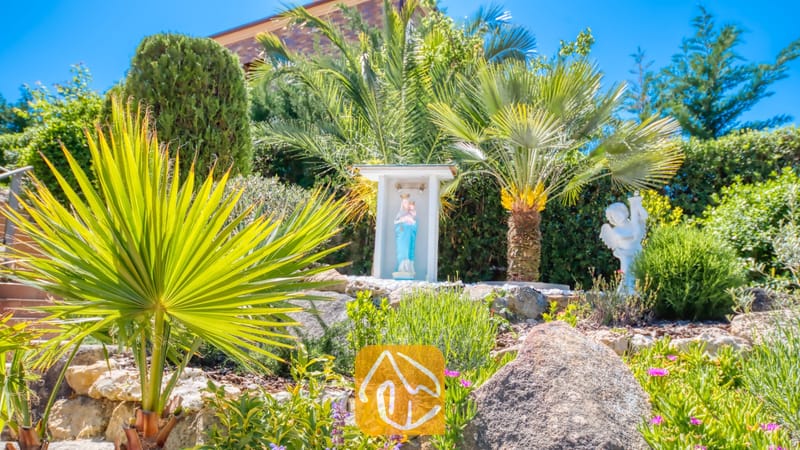 Vakantievilla's Costa Brava Spanje - Villa Jaruco - Zonnige tuin bij Villa Jaruco, voorzien van een klein kapelletje met een standbeeld, palmbomen en groene heesters op een rotstuinhelling.