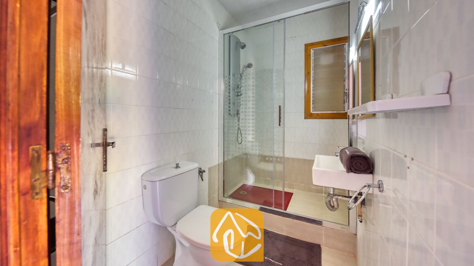 Vakantievilla's Costa Brava Spanje - Villa Jaruco - Wit betegeld badkamer met een glazen inloopdouche, een wit toilet en een compacte wastafel met wandbevestiging, voorzien van een opgerolde bruine handdoek.