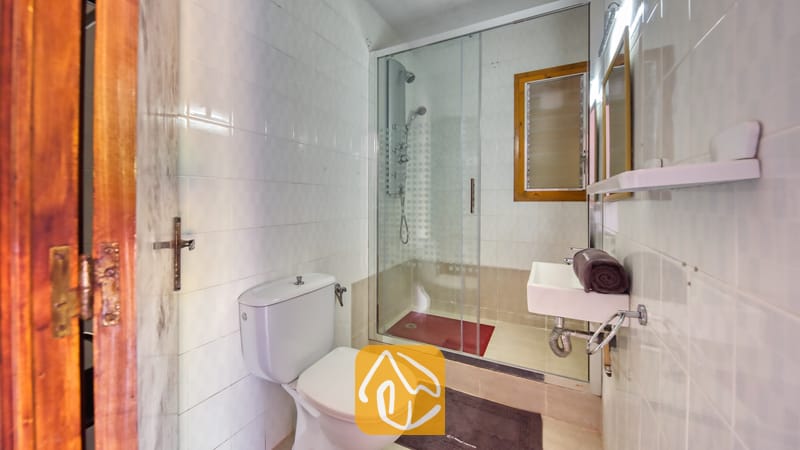 Villas de vacances Costa Brava Espagne - Villa Jaruco - Salle de bain carrelée de blanc comprenant une douche à l'italienne avec paroi en verre, des toilettes blanches et un lavabo compact suspendu accompagné d'une serviette marron roulée.