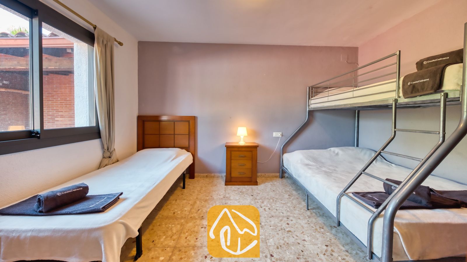 Vakantievilla's Costa Brava Spanje - Villa Jaruco - Slaapkamer met een houten eenpersoonsbed, een metalen stapelbed en een klein nachtkastje met een verlichte lamp daartussen, onder een raam met gordijnen.