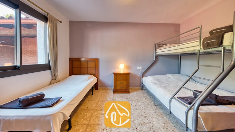 Villas de vacances Costa Brava Espagne - Villa Jaruco - Chambre avec un lit simple en bois, un lit superposé en métal et une petite table de chevet avec une lampe allumée entre les deux, sous une fenêtre avec des rideaux.