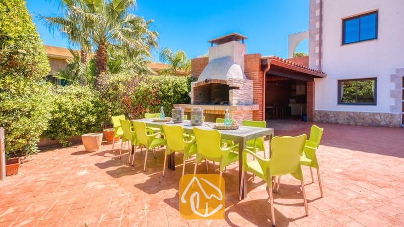 Vakantievilla's Costa Brava Spanje - Villa Jaruco - Eethoek in de buitenlucht bij Villa Jaruco, voorzien van een lange tafel met heldergroene stoelen, een ingebouwde stenen barbecue en omringende palmbomen.