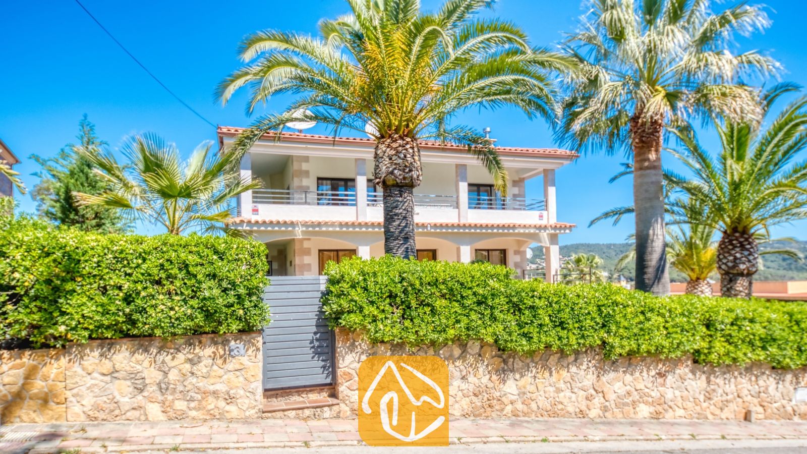 Vakantievilla's Costa Brava Spanje - Villa Jaruco - Buitengevel van Villa Jaruco met een wit gebouw van twee verdiepingen, hoge palmbomen en een stenen muur met een groene haag.