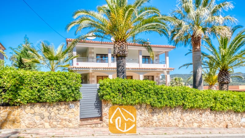 Villas de vacances Costa Brava Espagne - Villa Jaruco - Façade extérieure de la Villa Jaruco présentant un bâtiment blanc à deux étages, de grands palmiers et un mur en pierre surmonté d'une haie verte.