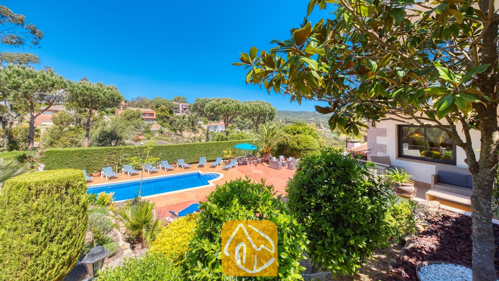 Vakantievilla's Costa Brava Spanje - Villa Jaruco - Het buitenterras van Villa Jaruco met een blauw zwembad, omzoomd door gestreepte ligbedden en omringd door groene bomen onder een helderblauwe hemel