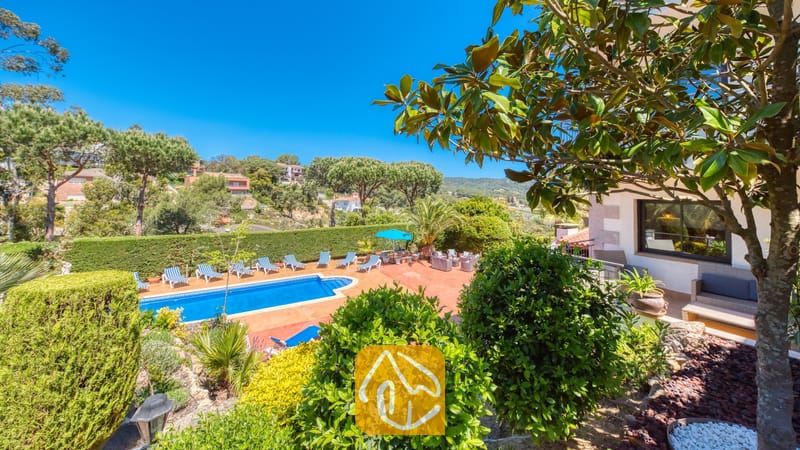 Vakantievilla's Costa Brava Spanje - Villa Jaruco - Het buitenterras van Villa Jaruco met een blauw zwembad, omzoomd door gestreepte ligbedden en omringd door groene bomen onder een helderblauwe hemel